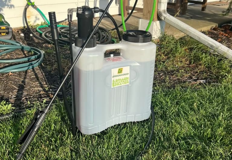 Translucent 4-gallon manual backpack sprayer resting on lawn grass next to garden hoses
