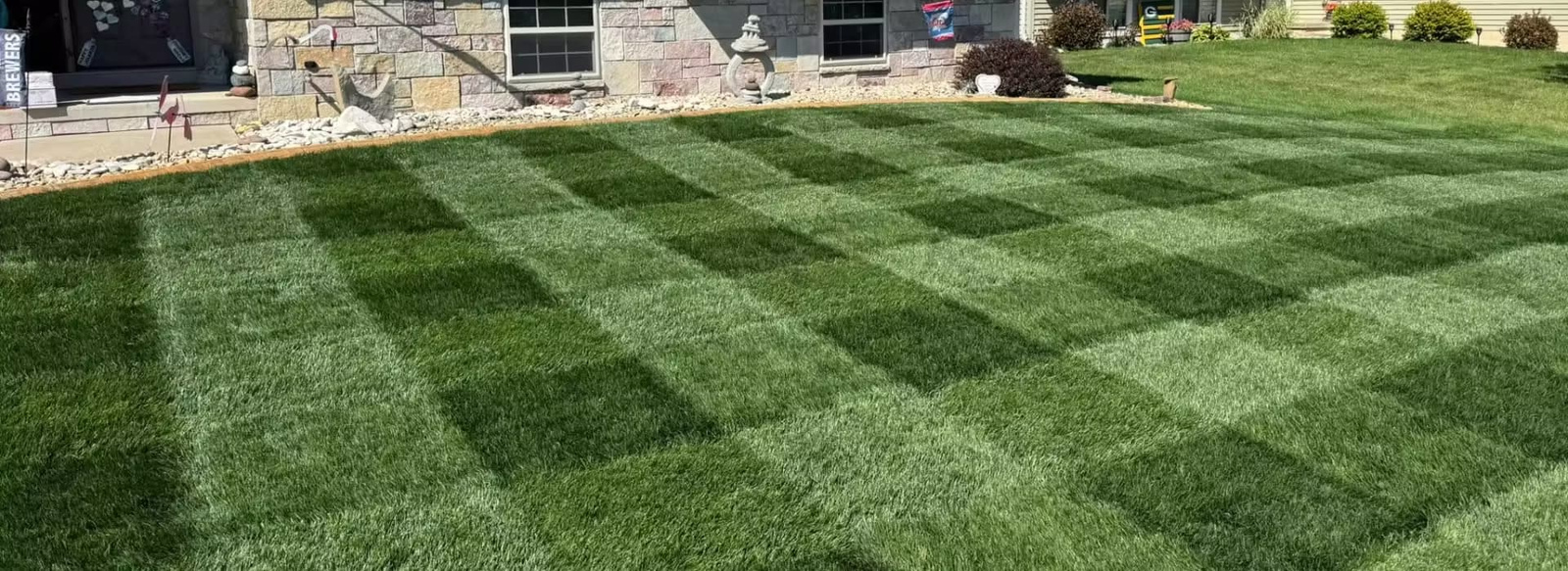 banner-contact A professionally manicured green lawn featuring a distinct checkerboard striped pattern achieved through cross-directional mowing.