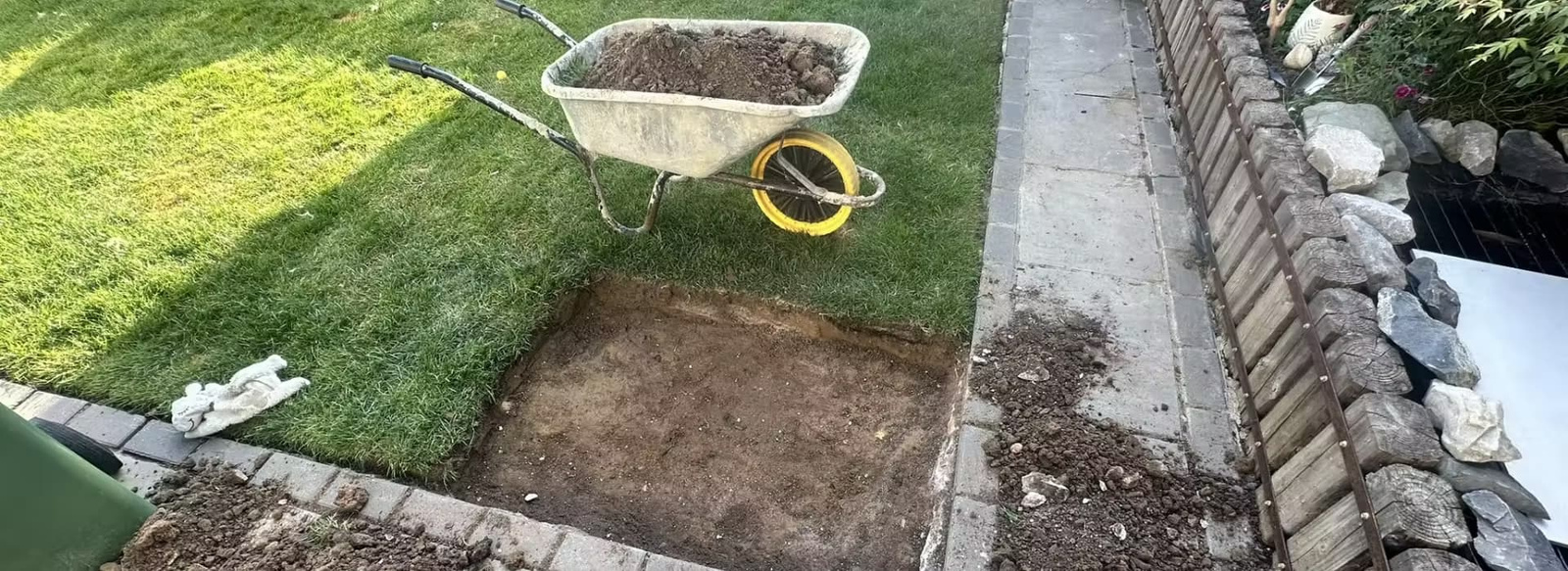 A deep rectangular excavation in a home lawn next to a wheelbarrow full of soil, documenting a structural substrate renovation for an MFY Field Story.