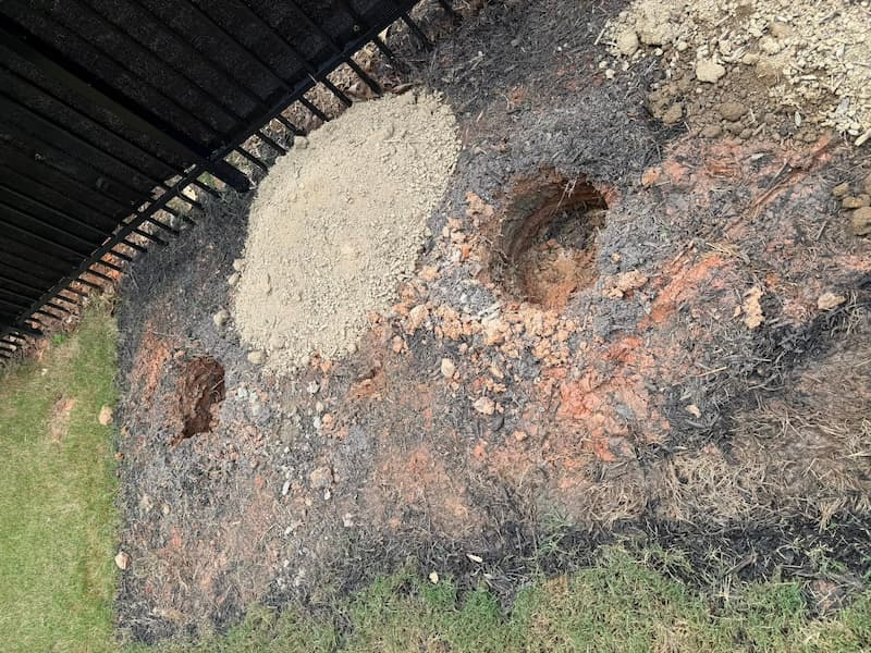 Two narrow planting holes dug in red clay soil next to a fence line showing slick glazed side walls