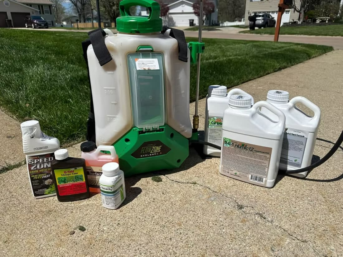 A battery-powered backpack sprayer standing on a suburban driveway beside bottles of lawn herbicide concentrate and surfactant, set up and ready for a weed-control application.