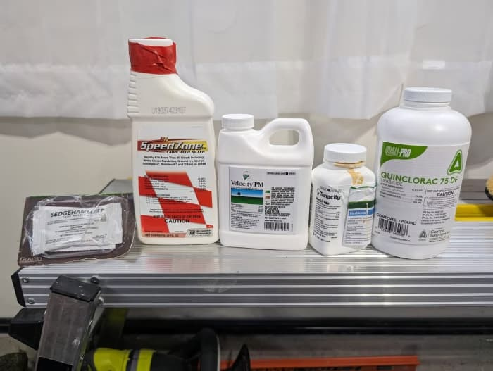 Five residential lawn herbicide containers lined up on a workbench — SedgeHammer, SpeedZone, Velocity PM, Tenacity, and Quinclorac 75 DF — each with a different active ingredient but all marked with the CAUTION signal word.
