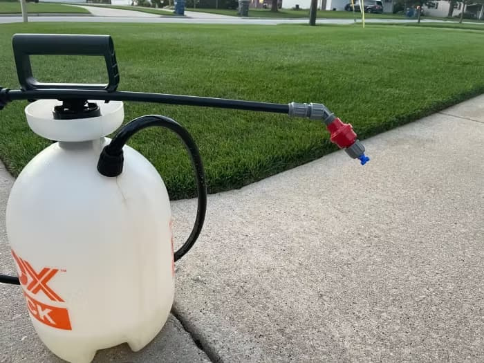 A white compression pump sprayer and wand sitting on a concrete path beside a well-maintained green lawn.