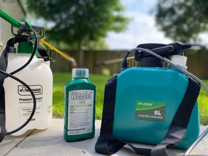 A Kingfisher hand-pump pressure sprayer beside a Sylstar 8-litre battery-powered sprayer, with a bottle of Weedol lawn weedkiller labeled "kills weeds not lawns" between them.