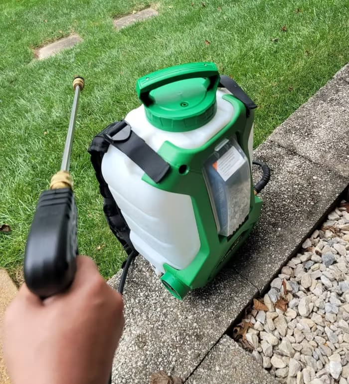 A person spraying lawn herbicide from a backpack sprayer while holding the wand with a bare hand and no long sleeve, showing missing personal protective equipment.