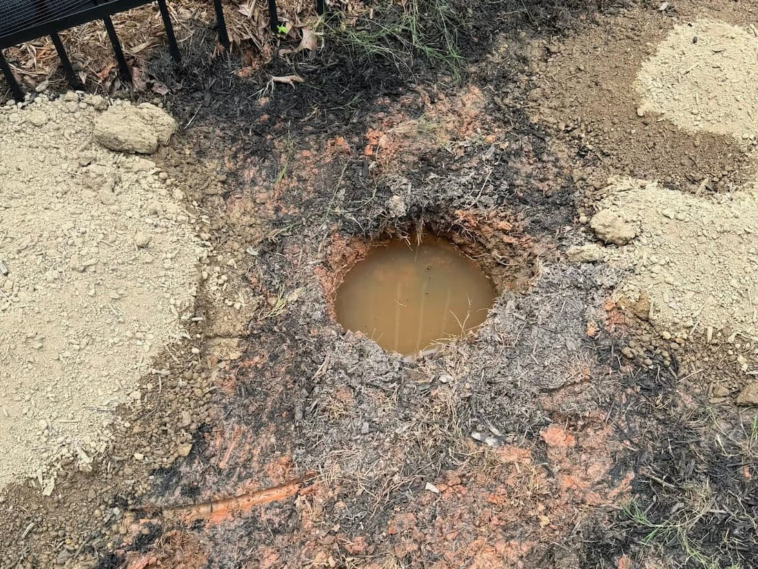 Overhead view of a narrow hole in heavy red clay soil completely filled with standing muddy water next to excavated soil piles