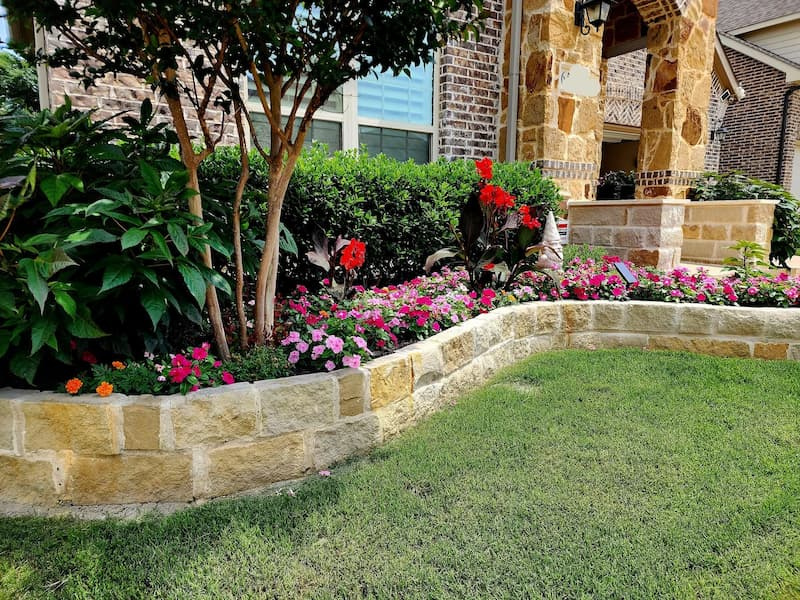 flower-bed-with-stone-border-along-lawn Red and pink flowering annuals in a landscaped bed with a stone retaining wall bordering a manicured lawn.