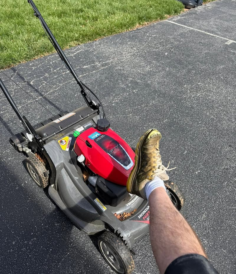 push-mower-on-driveway-with-foot-on-deck Push mower resting on a driveway with a foot on the mower deck.