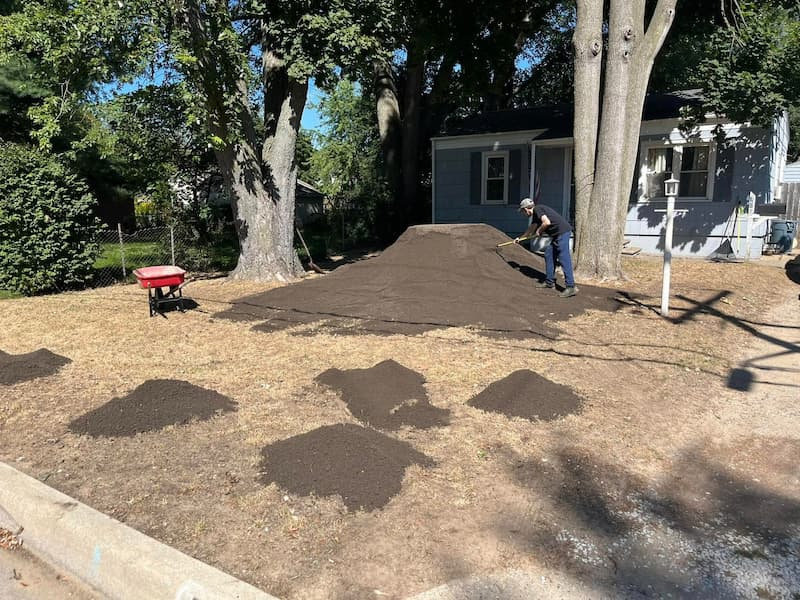 spreading-and-leveling-topsoil-with-rake Person raking and leveling mounds of dark topsoil in a backyard with a red wheelbarrow nearby.